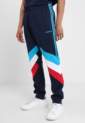 Person trägt marineblaue Adidas-Trainingshose mit türkis-, weiß- und rotfarbenen Chevron-Streifen, kombiniert mit weißen Turnschuhen, und steht vor einem schlichten Hintergrund.