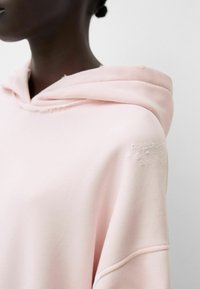 Světle růžová mikina s texturovaným pocitem, s volným střihem, připojenou kapucí a opotřebovanými detaily na rameni a rukávu.