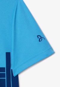 Kortärmad sporttröja i ljusblått tyg med en marinblå grafisk design och logo på vänster axel. Slät textur, fukttransporterande material.