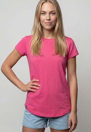 Junge Frau mit langen blonden Haaren, die ein leuchtend pinkes T-Shirt und hellblaue Jeansshorts trägt und mit einer Hand in der Hüfte steht.