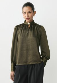 Oliivroheline satiinblouse, millel on kõrge krae, võtmeaugu dekoltee ja puhvis varrukad. Omab volditud detaile ja nööbitavaid mansette.