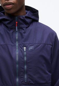 Muž nosiaci tmavomodrú kapucňovú bundu s prednými a zvislými vreckami na zips na hrudi a malým logom na ľavej strane hrude.