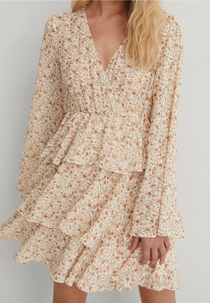 Femme portant une robe longue à manches longues couleur crème à motifs floraux, avec des volants superposés et un décolleté en V, debout devant un fond clair uni.