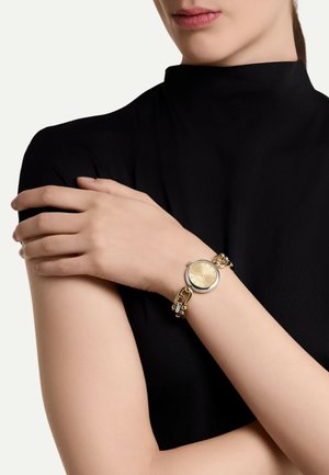 Goldfarbene Uhr mit rundem Zifferblatt, strukturiertem goldenen Ziffernblatt, silberfarbener Lünette und Gliederarmband; getragen am Handgelenk mit heller Hautfarbe.