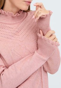 Pull en tricot rose avec col roulé côtelé et bordure froncée. Il présente des motifs texturés et des manches ajustées avec des poignets allongés.