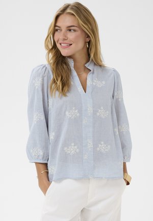 Žena s dlhými blond vlasmi, oblečená v svetlomodrej blúzke s bielou kvetinovou výšivkou a bielych nohaviciach, usmievajúca sa a pozerajúca doľava.