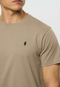 Vyras dėvi smėlinės spalvos trumpomis rankovėmis marškinėlį su mažos tamsiai mėlynos spalvos einančio vyro logotipu ant kairės krūtinės.