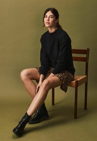 Un hanorac negru cu mâneci lungi, combinat cu o fustă din material leopard în nuanțe de maro. Subiectul poartă cizme negre, groase și stă pe un scaun din lemn.