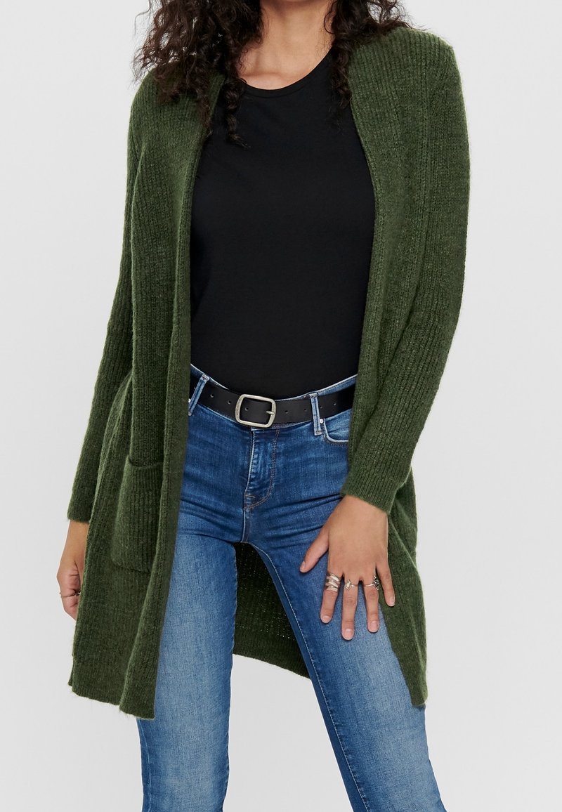 Femme portant un cardigan en maille vert foncé sur une chemise noire avec un jean bleu et une ceinture noire, main reposant sur la cuisse montrant plusieurs bagues.