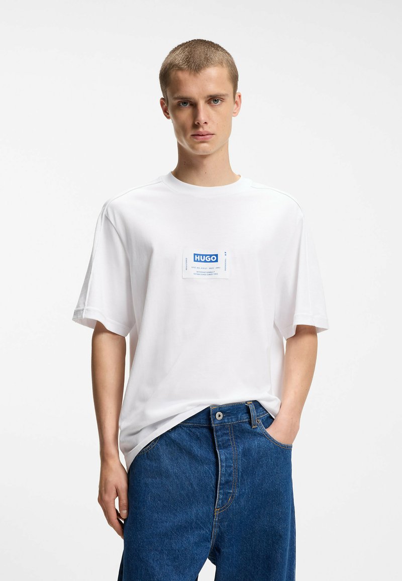 Jeune homme portant un t-shirt blanc avec un patch logo Hugo bleu et un jean bleu ample, debout avec une main dans la poche devant un fond uni.