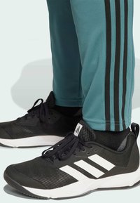 Zapatillas deportivas negras con parte superior de malla texturizada, rayas blancas en el costado y suela blanca, combinadas con pantalones deportivos verde azulado con rayas negras.