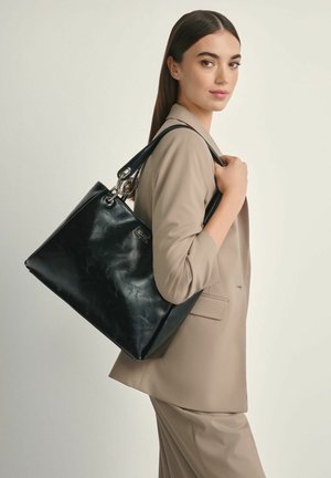 Kobieta w beżowym garniturze niosąca na ramieniu dużą czarną skórzaną torbę na tle jasnego tła.