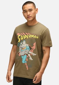 Camiseta de algodón verde oliva con un gráfico de Superman cargando a una mujer, con texto y elementos de diseño coloridos al estilo cómic.