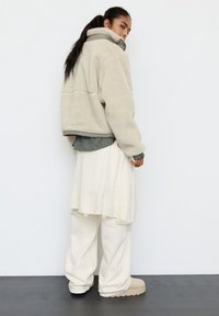 Fleecejakke i lys beige med høj krave, lagdelt over en grønternet skjorte, kombineret med hvide oversized bukser og beige støvler.