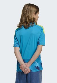 Turkos sporttröja med korta ärmar, med en gul detalj på axlarna och ett geometriskt blått mönster nära fållen.