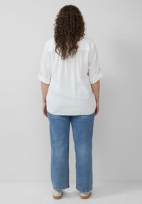 Witte, langarmige shirt met opgerolde mouwen, gecombineerd met lichtblauwe jeans en beige schoenen. Het haar is krullend en valt tot op de schouders.