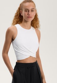 Bela brezrokavna majica s kratkim krojem in zasukanim sprednjim dizajnom, v kombinaciji z visokimi črnimi hlačami. Gladka tekstura, minimalističen slog.