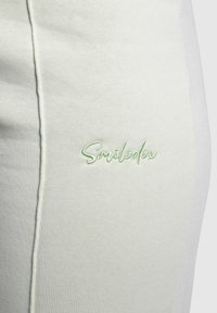 Vaaleanvihreä puuvillakangas, jossa on lähikuva brodeeratusta "Smiledax"-logosta, joka on ommeltu yhteensopivalla vihreällä langalla sivusauman varrella.