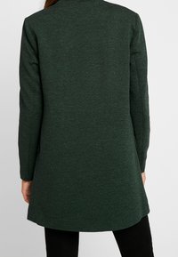 Manteau long texturé vert à manches longues, avec une silhouette droite et des poches latérales fendues, porté sur un pantalon noir.