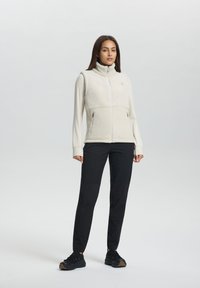Fleece-fodrad väst i krämfärgad med hög krage, dragkedjeslutning och två sidorfickor, parat med svarta byxor och svarta skor.