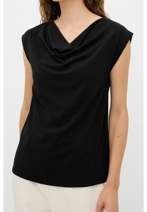 Woman wearing a sleeveless black top with a draped cowl neckline paired with light-colored pants against a plain background.