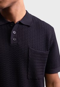 Laivastonsininen poolopaita, jossa on teksturoitu siksak-kuvio, vasemmalla rinnassa tasku ja kauluksen kolmen napin listallinen napitus.