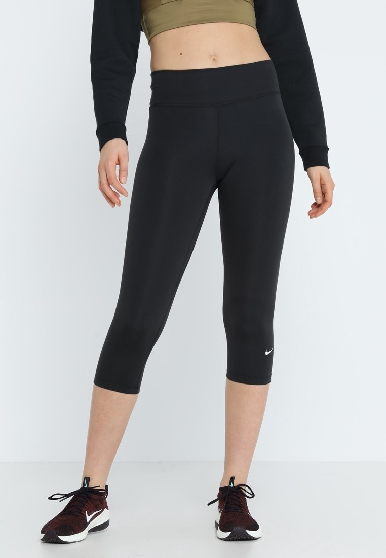 Personne portant des leggings courts noirs et des baskets Nike bordeaux, debout devant un fond blanc uni.