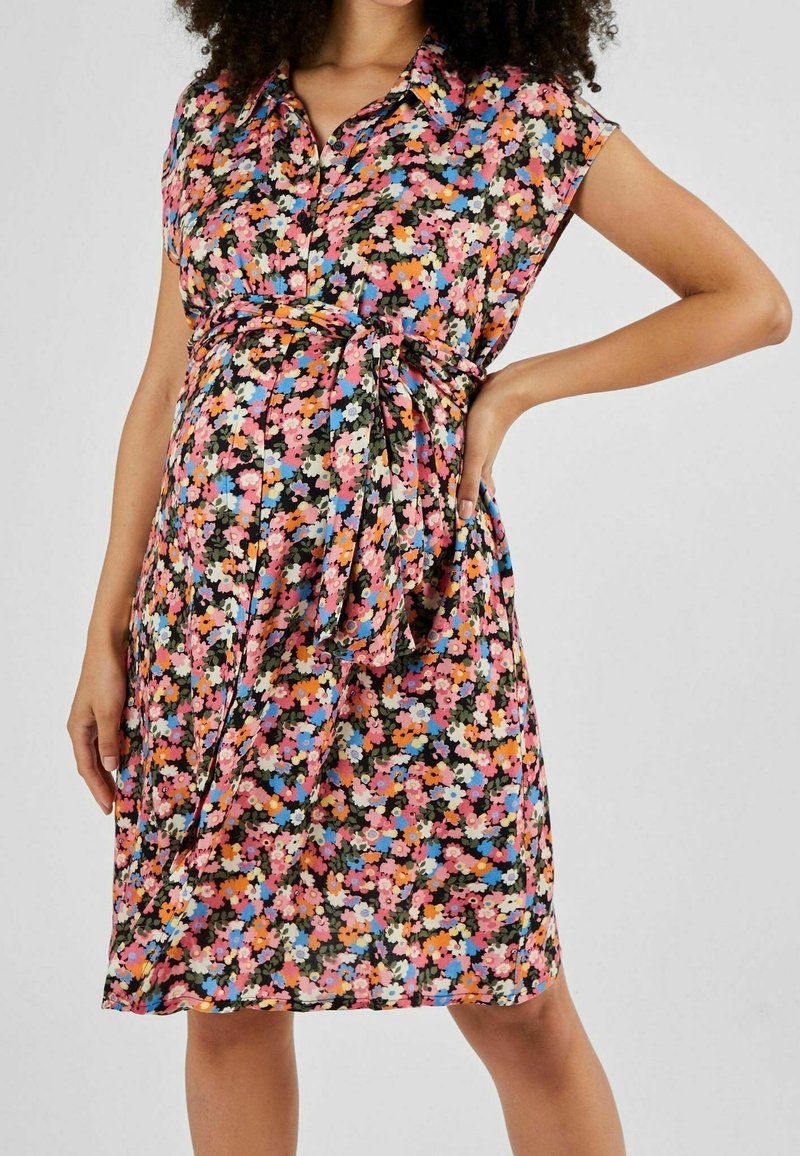 Femme enceinte portant une robe fleurie colorée longueur genou avec une ceinture à nouer, debout, une main sur la hanche et l’autre détendue.
