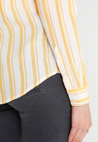 Camicia bianca con strisce verticali gialle, maniche lunghe, polsini abbottonati e orlo curvo. Indossata con jeans grigio scuro.