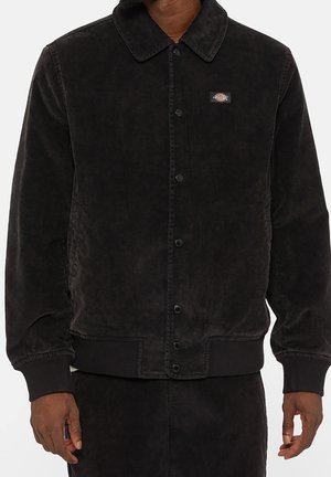 Schwarze Cordjacke zum Zuschließen mit Kragen und kleinem rechteckigem Logo-Patch auf der linken Brust, getragen von einer Person mit entspannten Händen.