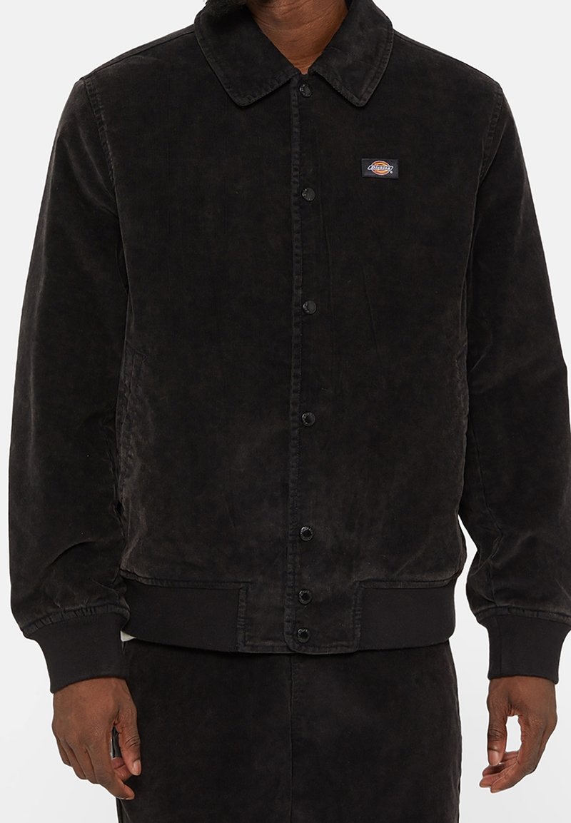 Veste noire en velours côtelé à boutons avec col et petit patch logo rectangulaire sur la poitrine gauche, portée par une personne aux mains détendues.