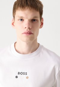 Valge puuvillane T-särk, millel on ümar kaelus, millel on sõna "BOSS" mustas kirjas kolme väikese värvilise ringi kohal, mille värvid on must, beez ja helepruun.