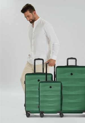 Mann in weißem Hemd und beiger Hose steht neben drei grünen Koffern mit ausgezogenen Griffen vor einfarbigem Hintergrund.