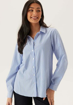 Kvinde med langt mørkt hår iført en blå og hvid ternet skjorte med knapper og sorte bukser, smilende og kigger til siden.