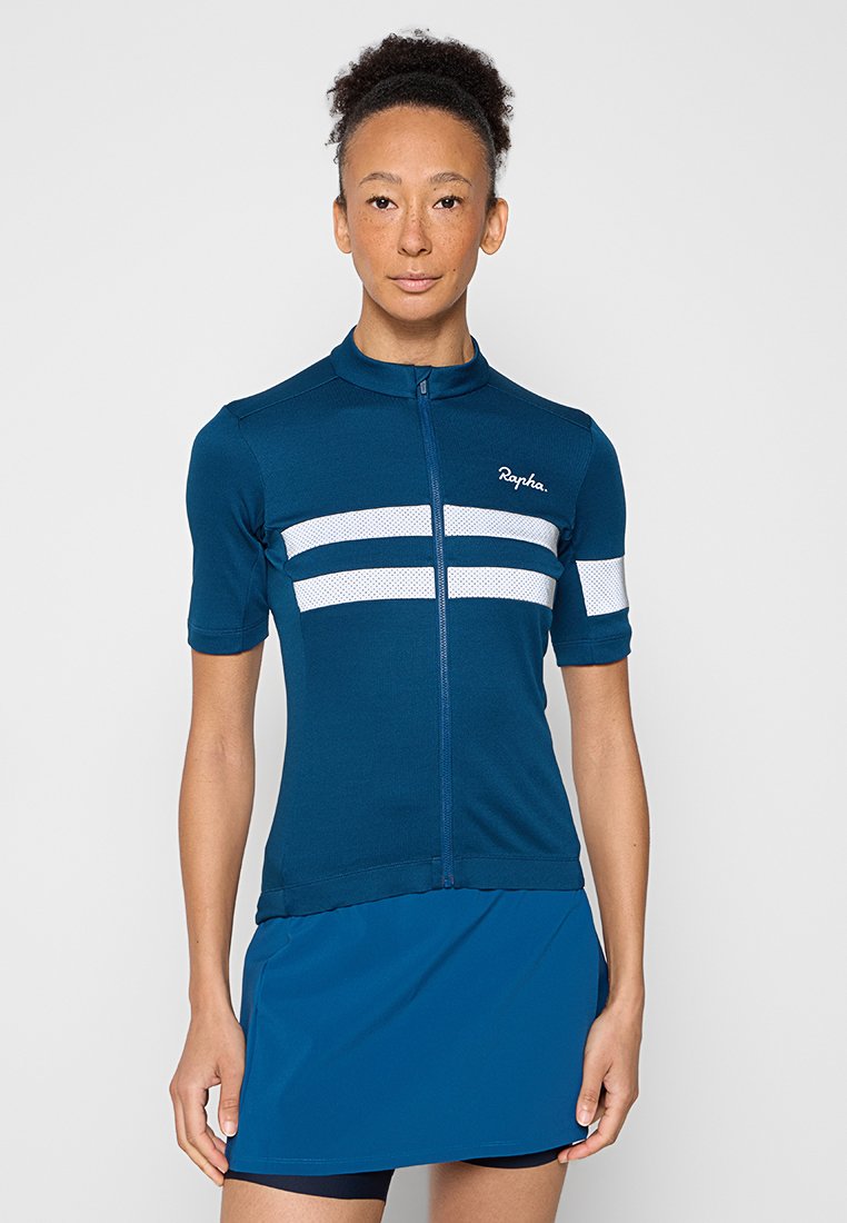 Vrouw in een turquoise fietsshirt met witte strepen en een bijpassende turquoise rok, staand tegen een effen achtergrond.