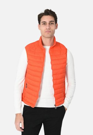 Uomo che indossa un gilet trapuntato arancione brillante senza maniche sopra una camicia bianca a maniche lunghe e pantaloni neri, in piedi con una mano in tasca.