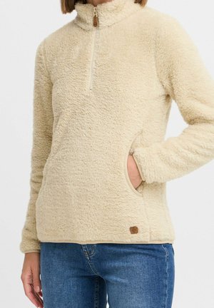 Person iført en beige fluffy fleecepullover med kort frontlynlås og hånd i sidelomme, kombineret med blå jeans.