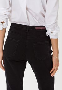 Pantalones de mezclilla negros con cintura alta, que cuentan con dos bolsillos traseros y una pequeña etiqueta de marca en la cintura. Textura suave.