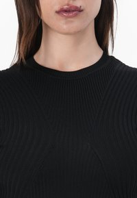 Juoda šonkauline megzta palaidinė su aukšta apykakle, tekstūruota medžiaga ir kampuotomis siūlėmis, kuri sukuria struktūruotą siluetą.