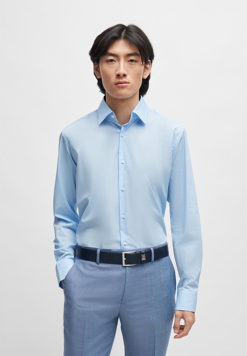 Camisa de mangas longas azul claro com colarinho abotoado, feita de um tecido suave, combinada com calças azul claro e um cinto de couro escuro.