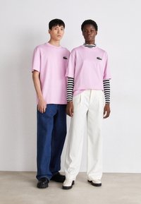 T-shirt di cotone rosa con un piccolo logo verde, abbinate a pantaloni in denim blu larghi e scarpe color crema, indossate da due modelli.