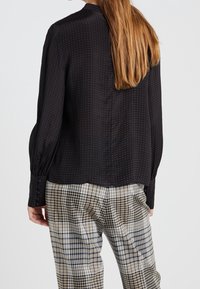 Svart blus med mönster, långa ärmar och rundad halsringning, kombinerad med ljusa rutiga byxor. Slät textur och avslappnad passform.