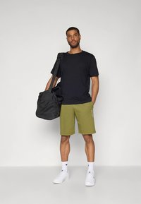 Fekete rövid ujjú póló, olívazöld rövidnadrág, fehér sportcipők és fekete sporttáska. A rövidnadrág oldalán logó részlet található.