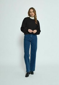 Črna pulover s balonskimi rokavi, v kombinaciji z modrimi jeans hlačami z visokim pasom. Manekenka nosi črne gležnjarje in stoji pred enostavno ozadjem.