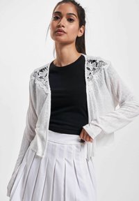 Weißer Cardigan mit Spitzenakzenten über einem schwarzen T-Shirt, kombiniert mit einem weißen plissierten Rock. Weicher Stoff mit offener Vorderseite und langen Ärmeln.