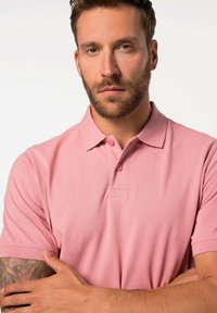 Rosa polotröja med kragdesign, korta ärmar och två knappar vid halsen. Tyget verkar mjukt med en lätt textur.