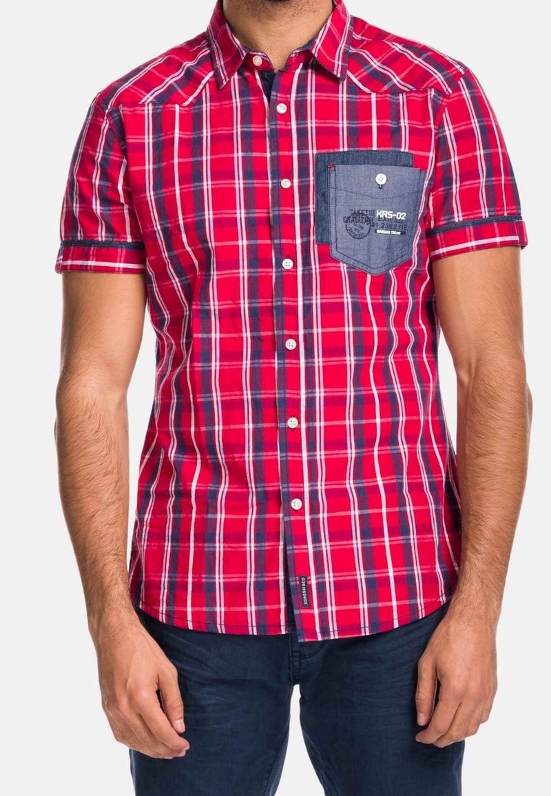 Homme portant une chemise à manches courtes à carreaux rouges avec une poche poitrine bleue et un pantalon bleu foncé, debout, les bras détendus.