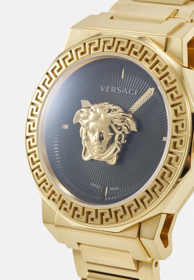 Gold-tone Versace watch featuring a black metallic face, a Medusa emblem at the center, and a patterned bezel with Greek key details.