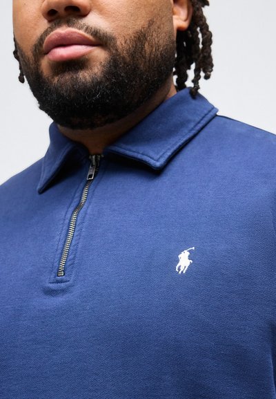 Άνδρας με γενειάδα που φοράει μπλε πόλο μπλούζα με φερμουάρ μέχρι το στήθος, με ένα μικρό λευκό κεντημένο λογότυπο παίκτη πόλο στο στήθος.