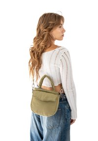 Olivovo zelená polstrovaná taška na rameno s okrúhlym uchom, s prednou vreckovou chlopňou, pripevnená k osobe v bielom rebrovanom svetri a džínsoch.